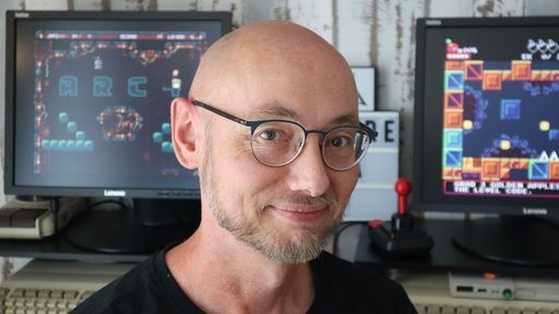 Hans-Gerhard Surey in seinem Arbeitszimmer. Er blick direkt in die Kamera. Er trägt eine Brille und einen kurzen Bart. Im Hintergrund laufen auf zwei Bildschirmen Videospiele.