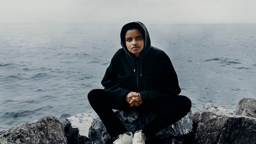 Schauspielerin hockt in schwarzem Hoodie vor Meer