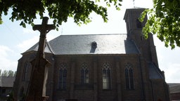 Die Kirche St. Johann Baptist in Sievernich bei Vettweiß bei Sonnenschein von der Seite und hinter Bäumen