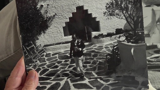 Ein Schwarz-Weiß-Foto zeigt Salvador Dali, wie er den Hof seines Hauses überquert, man sieht am Bildrand eine Hand, die das Foto hält.