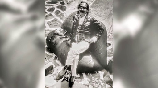 Ein Schwarz-Weiß-Foto zeigt den Künstler Salvador Dali auf einem Sofa sitzend, es sieht aus wie ein Kussmund