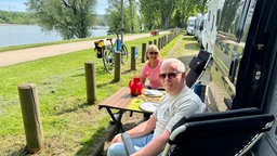 eine Frau und ein Mann sitzen bei sonnigem Wetter vor einem Camper am Flussufer