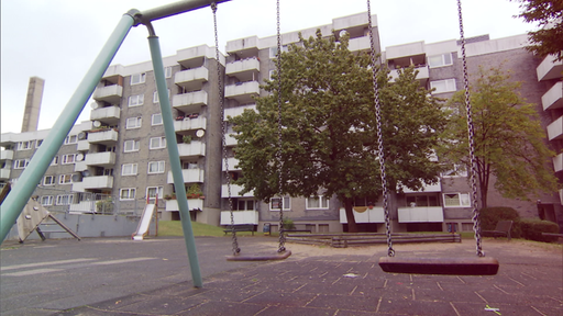 Zwei verwaiste Schaukeln an einem Spielplatz. Im Hintergrund ist ein mehrstöckiger Gebäudekomplex in dem sich die Wohnung der Täterin befindet. Der Plattenbau wirkt heruntergekommen.