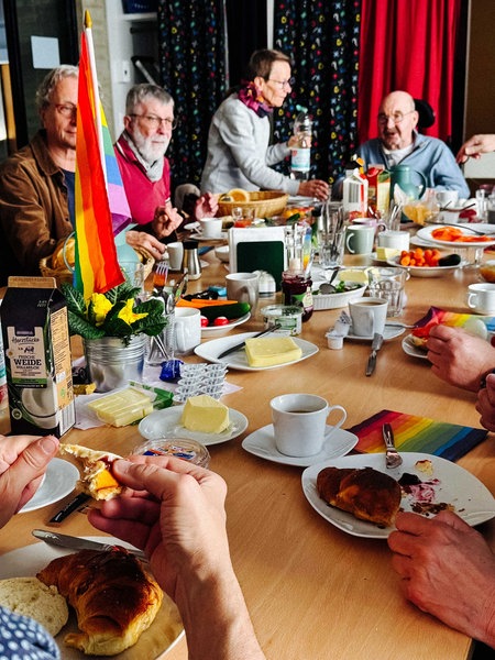 Senioren sitzen um einen gedeckten Frühstückstisch herum. Auf dem Tisch steht eine Regenbogenflagge.