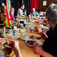 Senioren sitzen um einen gedeckten Frühstückstisch herum. Auf dem Tisch steht eine Regenbogenflagge.