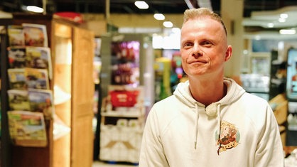 Ein Mann steht im Supermarkt. Er schaut leicht rechts an der Kamera vorbei und lächelt.