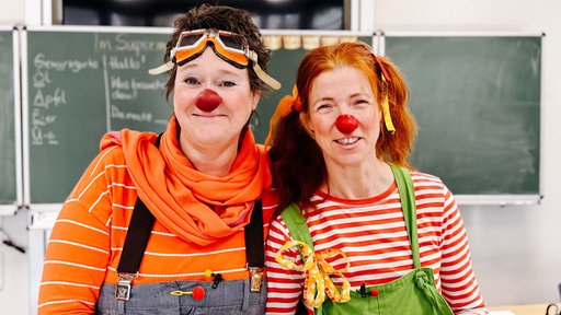 Zwei Frauen sind als Clowns kostümiert und stehen in einem Klassenraum vor einer Tafel.