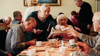 Mehrere Menschen an einer Kaffeetafel, eine Frau verteilt Zettel