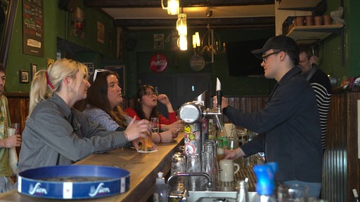 Frauen sitzen an einer Theke, ein Mann zapft dahinter ein Bier.
