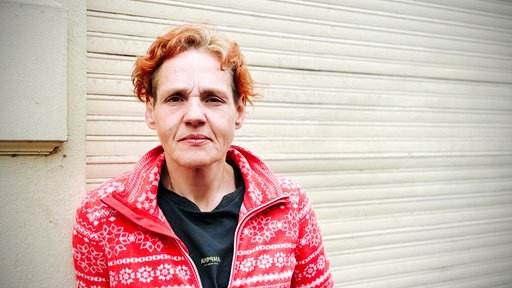 Susan steht in einem rot-weißen Pullover vor einer Wand und guckt in die Kamera