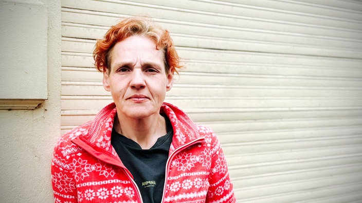 Susan steht in einem rot-weißen Pullover vor einer Wand und guckt in die Kamera