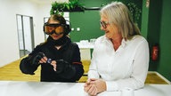 Eine blonde Frau trägt einen Altersanzug, bestehend aus einem beschwerten schwarzen Arbeitsanzug, Handschuhen und einer dunklen Skibrille, und versucht, ein Bonbon zu öffnen.