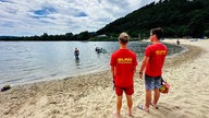 Henning Thielemeyer und Philipp Fögen stehen am Strand vom Godelheimer See und blicken auf den See, auf ihren roten T-Shirts steht auf dem Rücken "DLRG Wasserrettung"