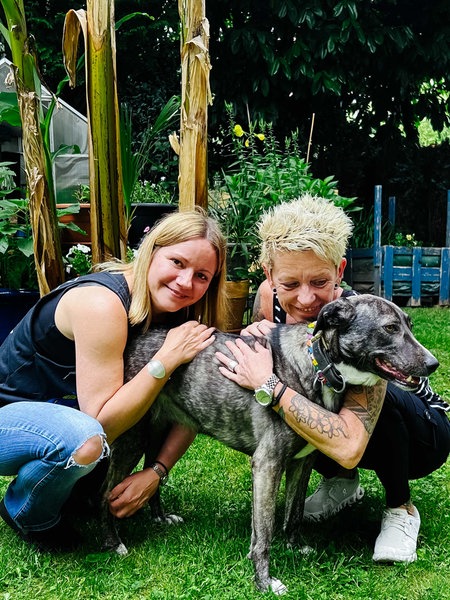Zwei Frauen, Nadin Duberg und Susi Schmücker, hocken in einem Garten und umarmen ihre grau-weiße Hündin Wally.