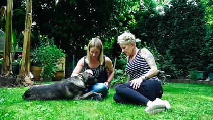 Nadin Duberg und Susi Schmücker sitzen auf dem Boden, auf Gras, Nadine Duberg streichelt mit einer Hand die Hündin Wally.
