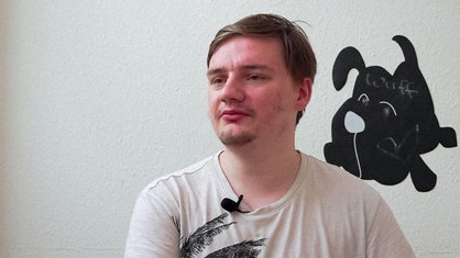 Ein junger Mann sitzt in einem Raum und schaut rechts an der Kamera vorbei.
