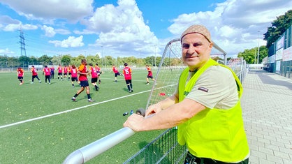 Suat Amaҫ in gelber Warnweste steht am Geländer neben dem Fußballplatz, auf dem Spieler im roten Trikot gerade trainieren.