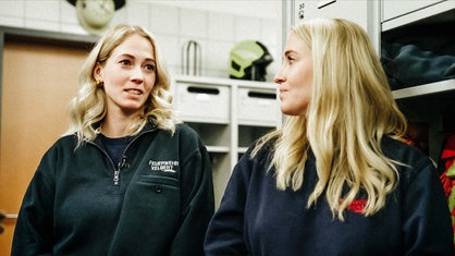 Zwei blonde Frauen stehen im Gerätehaus einer Feuerwache und sprechen miteinander