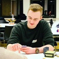 Jan Berkemeyer, ein junger blonder Mann im grünen Pullover, sitzt hinter einem weißen Tisch und schaut auf sein Smartphone, das er in den Händen hält.