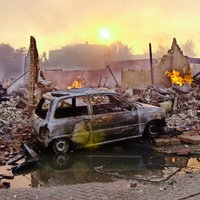 Ein ausgebranntes Auto steht vor Gebäudetrümmern. Im Hintergrund lodern weiter Flammen.