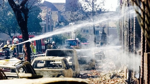 Feuerwehrleute löschen einen Gebäude, vor den viele ausgebrannte Autos stehen.