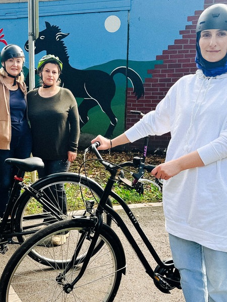 Drei Frauen mit Helm stehen neben zwei Fahrrädern auf der Straße.