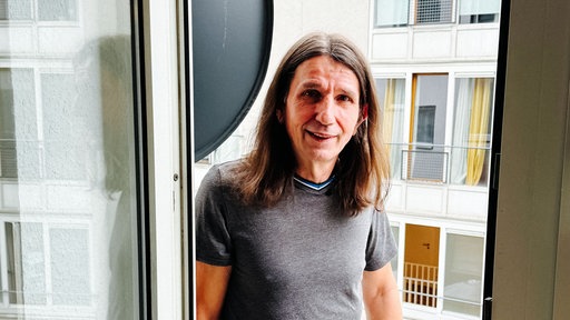 Willi Lehmann steht auf dem Balkon seiner Kölner Wohnung, lehnt am Türrahmen und lächelt.