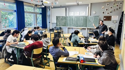 Der Unterricht der 10b der Green-Gesamtschule. Vorne an der Tafel steht ein Lehrer. Die Schülerinnen und Schüler sitzen verteilt an Tischgruppen. 