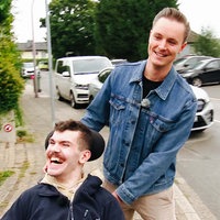 Hospizdienstmitarbeiter Jan Meyer schiebt Hubertus Kaiser in einem Rollstuhl über einen Bürgersteig.