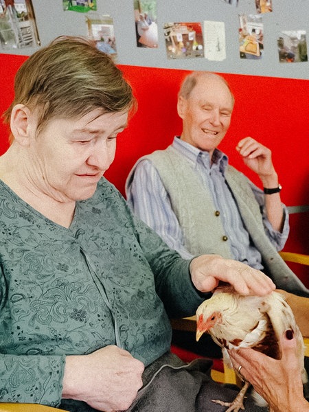 Birgit Brammertz hält ein Huhn. Neben ihr sitzt eine Seniorin, die das Huhn streichelt.