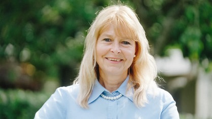 Birgit Brammertz in blauer Bluse sitzt auf einem Stuhl. Sie lächelt in die Kamera.