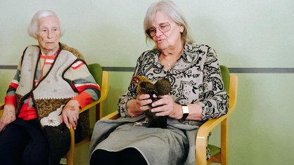 Eine Seniorin streichelt ein Huhn, das auf ihrem Schoß sitzt.