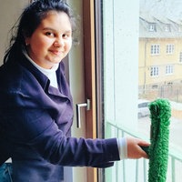 Gabriela, eine junge brünette Frau im schwarzen Pullover, steht vor einem Fenster und hält einen nassen grünen Wischer gegen die Scheibe.