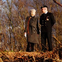 Ein Mann und eine Frau stehen nebeneinander an einem spätherbstlichen Wald und schauen sich an. 