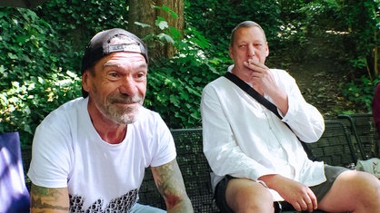 Ein Mann sitzt auf einer Bank und lächelt in die Kamera, neben ihm sitzt ein weiterer Mann, der raucht.