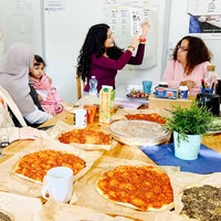 Bilge Akbulut hält einen Zettel hoch auf dem verschiedene Vokabeln erklärt werden. Die Teilnehmerin hören ihr interessiert zu. Auf dem Tsich stehen mehrere libanesische Pizzen.