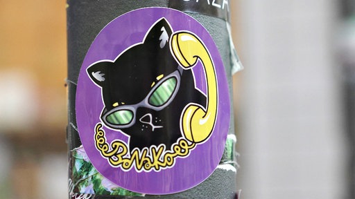 Ein runder Aufkleber mit dem Logo des Bonner Nachtkollektivs: Auf lila Hintergrund ist eine schwarze Katze mit Sonnenbrille und einem gelben Telefonhörer am Ohr zu sehen, die gelbe Schnur bildet den Schriftzug "BoNaKo".