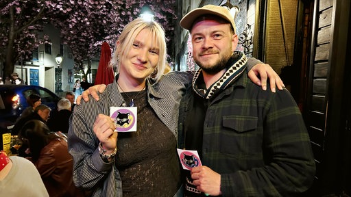 Neele Schumann, eine junge blonde Frau mit schulterlangen Haaren im gestreiften Hemd, steht links neben Stefan Lindner, einem bärtigen Mann mit weißer Kappe und dunklem Hemd. Sie halten quadratische Aufkleber in der Hand.