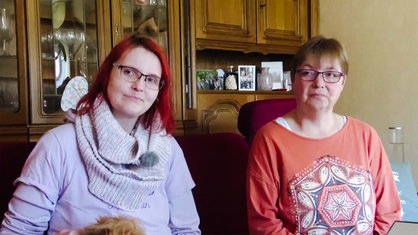 Annika Witteler sitzt mit ihrer Mutter auf einer Couch. Hinter ihnen steht ein Holzregal mit mehreren gerahmten Bildern.