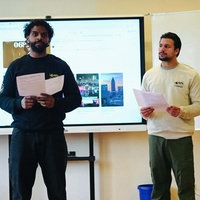 Zwei Männer, Kevin Grund und Nigash Sriranganathan, stehen mit Zetteln in der Hand in einer Schulklasse, hinter ihnen ist eine Präsentation zu sehen.