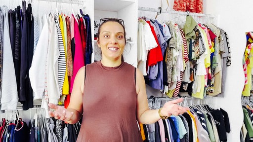 Sevil Mokhtare mit braunem, ärmellosen Kleid, Sonnenbrille auf dem Kopf, Haare im Zopf zurückgebunden, lächelt, beide Hände geöffnet und auf Brusthöhe gehoben, im Hintergrund hängen Klamotten