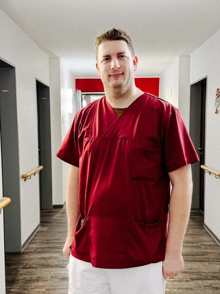 Robbin Nilles steht in einem Gang. Er trägt einen Kasack und lächelt.