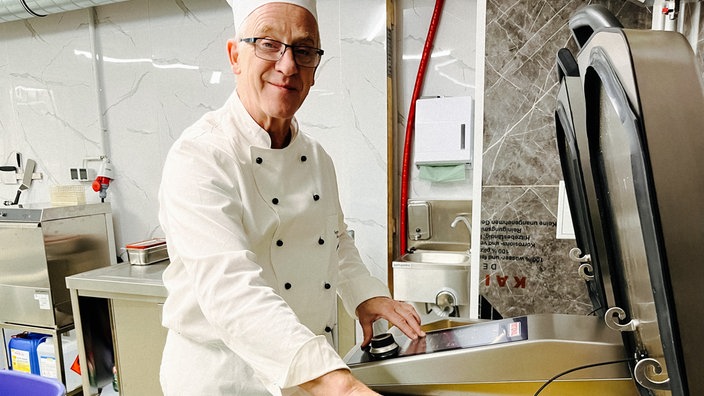 Klaus Wellmeier in seiner Suppenküche. Seine Hand an einem großen Behälter, in dem ein Eintopf zu sehen ist.