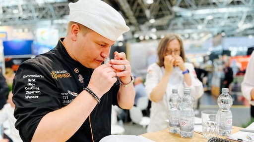 Patrick Zimmer riecht an einem kleinen Gläschen. Im Hintergrund steht eine Frau, die auch an einem Gläschen riecht.