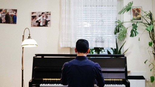 Aeham Ahmad sitzt an seinem Klavier in seiner Wohnung. An der Wand hängen Bilder. Neben dem Klavier ist eine Stehlampe, so wie eine Pflanze zu sehen.