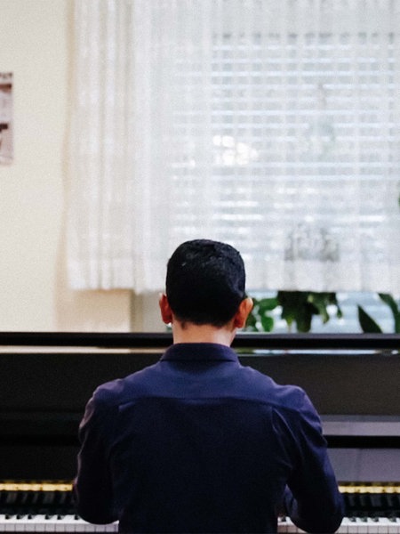 Aeham Ahmad sitzt an seinem Klavier in seiner Wohnung. An der Wand hängen Bilder. Neben dem Klavier ist eine Stehlampe, so wie eine Pflanze zu sehen.