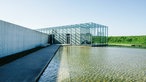Ein langer Glasbau, der auf einen künstlich angelegten Teich ragt.