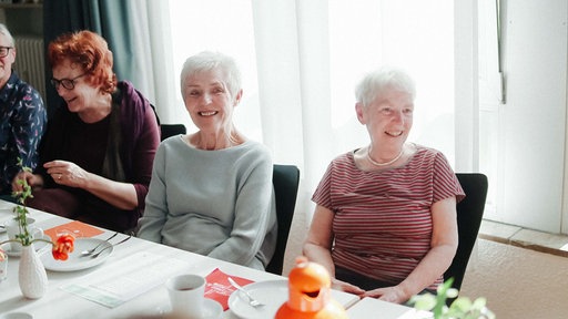 Ein gedeckter Tisch. Vier Frauen sitzen daran. Sie lachen.