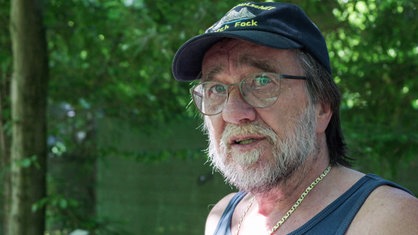 Klaus Mindrup mit grauem Bart und Brille steht im Grünen. Er trägt eine blaue Kappe und hat eine goldene Kette um den Hals. 