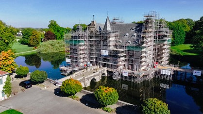 Das eingerüstete Schloss Bodelschwingh, von einem Wassergraben umgeben vor bewaldeter Landschaft.